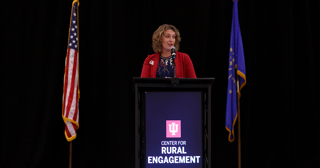 woman with curly hair standing in front of a mic and podium with American flag on left and a blue flag on the right