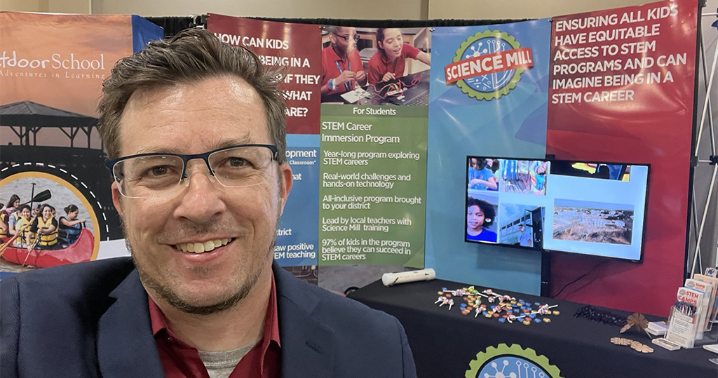 man wearing glasses a red collared shirt and blazer standing in front of posters and a computer screen